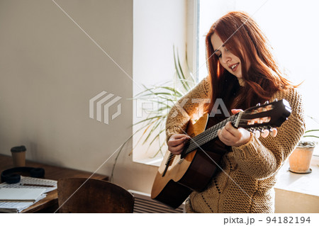 Indoor cozy Hobbies for winter, autumn cold season. Redhead woman playing acoustic guitar and listening to music, sitting near window home Indoor cozy Hobbies for winter, autumn cold season. Redhead woman playing acoustic guitar and listening to music, sitting near window home 94182194