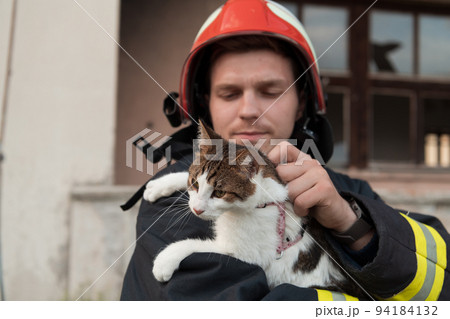 Close-up portrait of heroic fireman in protective suit and red helmet holds saved cat in his arms. Firefighter in fire fighting operation. Close-up portrait of heroic fireman in protective suit and red helmet holds saved cat in his arms. Firefighter in fire fighting operation. 94184132