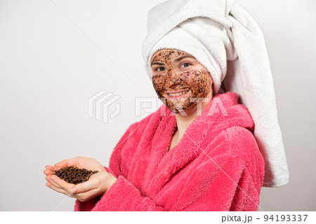 A woman in a pink bathrobe with a coffee mask on her face holds coffee beans in her hands A woman in a pink bathrobe with a coffee mask on her face holds coffee beans in her hands 94193337