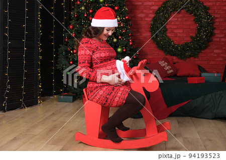 A pregnant woman prepares for Christmas, sits on a red children's wooden horse and puts a hat on her belly. A pregnant woman prepares for Christmas, sits on a red children's wooden horse and puts a hat on her belly. 94193523