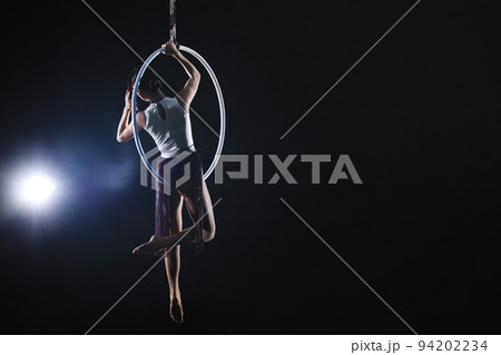 Young woman performing acrobatic element on aerial ring against dark background. Space for text 94202234
