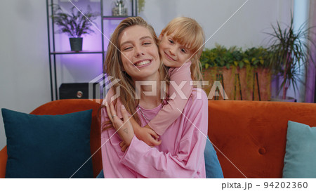 Smiling child kid daughter embrace young mother, look at camera, sweet tender love at home sofa Smiling child kid daughter embrace young mother, look at camera, sweet tender love at home sofa 94202360
