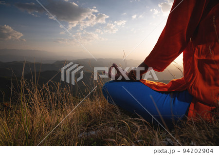 Close-up of a woman and female hands sitting in a lotus position, yoga outdoors in the grass at sunset against the backdrop of a mountain range and clouds, meditation Close-up of a woman and female hands sitting in a lotus position, yoga outdoors in the grass at sunset against the backdrop of a mountain range and clouds, meditation 94202904