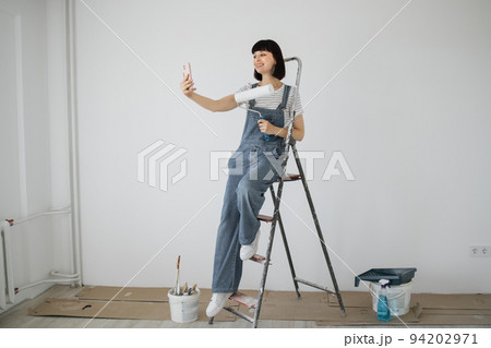 Red hair girl with cute smile holds a paint roller and smart phone in her hands. Red hair girl with cute smile holds a paint roller and smart phone in her hands. 94202971