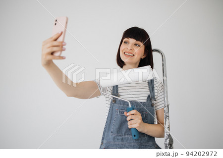 Red hair girl with cute smile holds a paint roller and smart phone in her hands. Red hair girl with cute smile holds a paint roller and smart phone in her hands. 94202972