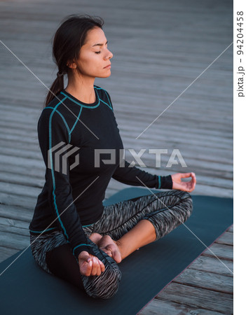 Concentrated girl in black practicing in yoga meditation on wooden seafront. Woman in lotus pose 94204458