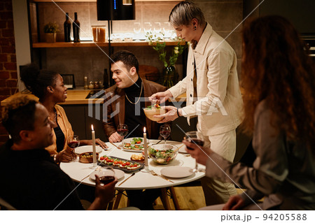 Young man putting vegetable salad on his plate while standing by dinner table 94205588