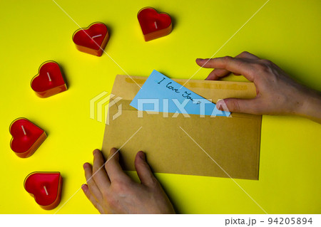 A woman's hand puts a blue note in a cardboard envelope with a handwritten text I LOVE YOU for her lover on Valentine's Day. Love letter on a yellow background flatly. Red candles in shape of hearts. 94205894