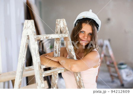 Young attractive girl in beige dress and protective helmet poses on stepladder in room being renovated 94212609