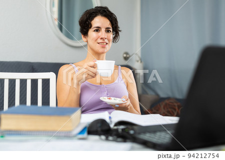 Woman drinking coffee in front of laptop while having video call conversation 94212754