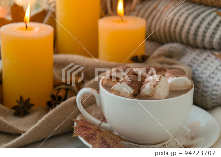 Celebrating autumn holidays at cozy home with cup hot chocolate marshmallows cacao on the windowsill Hygge atmosphere Thanksgiving candle on cozy knitted sweater in warm yellow lights 94223707