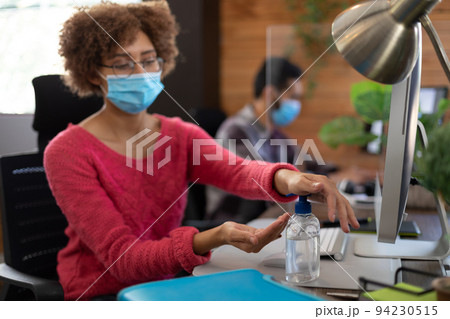 Mixed race woman wearing face mask sanitizing hands at desk in office 94230515