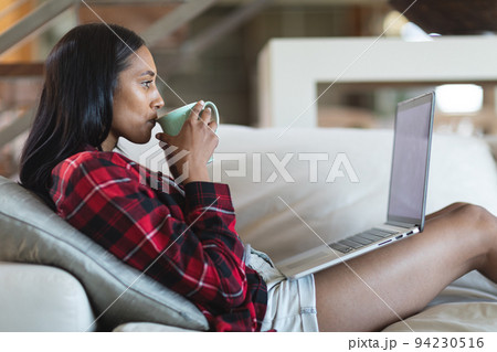 Mixed race woman drinking coffee using laptop computer at home 94230516