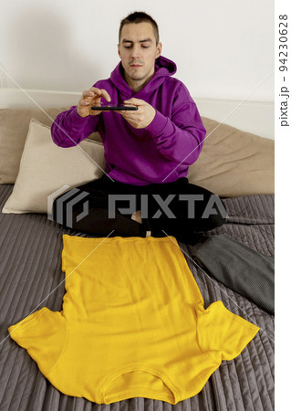Man with violet hoodie using digital tablet, takes a photo of his old clothes to sell them online. Selling on website, e-commerce. Reuse, second-hand concept. Conscious consumer, sustainable lifestyle Man with violet hoodie using digital tablet, takes a photo of his old clothes to sell them online. Selling on website, e-commerce. Reuse, second-hand concept. Conscious consumer, sustainable lifestyle 94230628