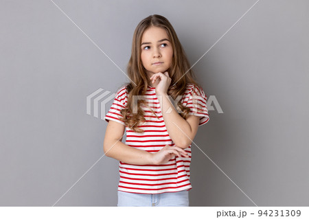 Portrait of thoughtful pensive little dark haired girl wearing striped T-shirt standing holding chin, thinks about something important. Indoor studio shot isolated on gray background. Portrait of thoughtful pensive little dark haired girl wearing striped T-shirt standing holding chin, thinks about something important. Indoor studio shot isolated on gray background. 94231309