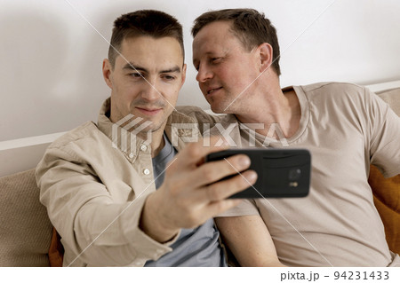 Happy gay couple with casual clothes spending time together at home and making selfie on smartphone. Homosexual relationships and alternative love. Cosy interior. 94231433