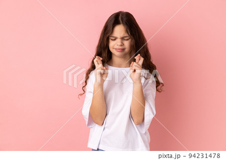 Portrait of little girl wearing white T-shirt crossing fingers in hope for good luck, making wish, rituals, standing with closed eyes. Indoor studio shot isolated on pink background. Portrait of little girl wearing white T-shirt crossing fingers in hope for good luck, making wish, rituals, standing with closed eyes. Indoor studio shot isolated on pink background. 94231478