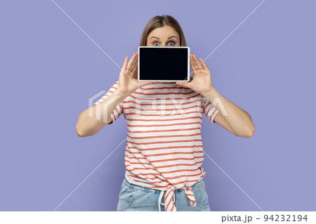 Portrait of shocked astonished blond woman wearing striped T-shirt covering half of face with tablet, showing empty display with copy space. Indoor studio shot isolated on purple background. Portrait of shocked astonished blond woman wearing striped T-shirt covering half of face with tablet, showing empty display with copy space. Indoor studio shot isolated on purple background. 94232194