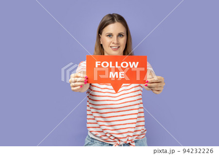 Portrait of joyful beautiful blond woman wearing striped T-shirt holding out red card with follow me inscription, asking to subscribe to her blog. Indoor studio shot isolated on purple background. Portrait of joyful beautiful blond woman wearing striped T-shirt holding out red card with follow me inscription, asking to subscribe to her blog. Indoor studio shot isolated on purple background. 94232226