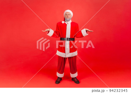 Full length portrait of elderly man with gray beard wearing santa claus costume shrugging shoulders as doesn't know answer, being uncertain, not sure. Indoor studio shot isolated on red background. Full length portrait of elderly man with gray beard wearing santa claus costume shrugging shoulders as doesn't know answer, being uncertain, not sure. Indoor studio shot isolated on red background. 94232345