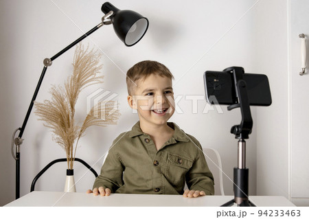 Adorable, cute, little boy blogger recording lifestyle blog, talking to camera of smartphone on tripod. Young influencer filming vlog for his channel. Child makes video for his followers online. 94233463