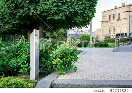 modern iron lantern of ground garden led lighting with bushes in city street with trees near flower bed landscape next to the ramp and steel railings, in the background is an old town house, nobody. 94235329