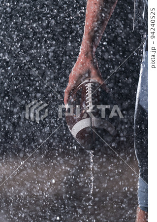 Close up of American Football Athlete Warrior Standing on a Field focus on ball and Ready to Play. Player Preparing to Run, Attack and Score Touchdown. Rainy Night with Dramatic lens flare and rain Close up of American Football Athlete Warrior Standing on a Field focus on ball and Ready to Play. Player Preparing to Run, Attack and Score Touchdown. Rainy Night with Dramatic lens flare and rain 94240525