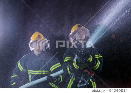Firefighters using a water hose to eliminate a fire hazard. Team of female and male firemen in dangerous rescue mission. Firefighters using a water hose to eliminate a fire hazard. Team of female and male firemen in dangerous rescue mission. 94240814