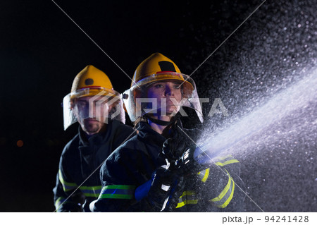 Firefighters using a water hose to eliminate a fire hazard. Team of female and male firemen in dangerous rescue mission.  Firefighters using a water hose to eliminate a fire hazard. Team of female and male firemen in dangerous rescue mission.  94241428