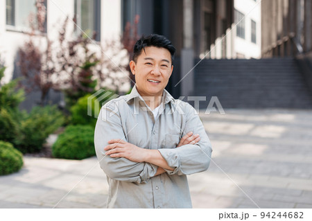 Portrait of confident asian entrepreneur posing with crossed arms and looking at camera near office building 94244682