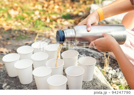 Close-up top above view woman hand hold steel thermos pouring hot tea many white paper cup group picnic mountain trekking outdoors fall sunny morning evening. Person thermo flask warm drink sharing 94247790
