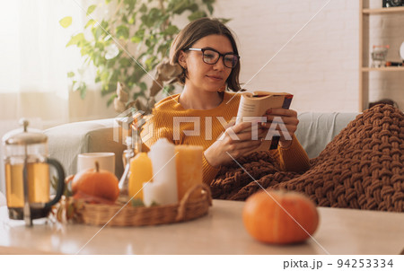 Lady reads a book in blanket on the sofa in front of teapot and candles 94253334