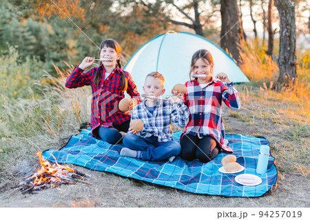 A cheerful company of two girls and a boy on a picnic in the middle of the forest. Children fry sausages on the fire, eat buns and have fun in nature. The concept of active recreation in the summer A cheerful company of two girls and a boy on a picnic in the middle of the forest. Children fry sausages on the fire, eat buns and have fun in nature. The concept of active recreation in the summer 94257019
