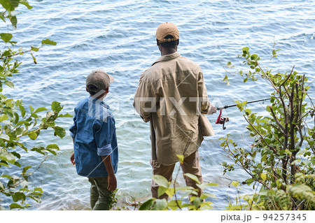 Rear view of senior black man with rod and his grandson standing by lake Rear view of senior black man with rod and his grandson standing by lake 94257345