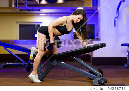 Portrait of young beautiful athletic fitness athlete woman doing exercises in gym with dumbbells, looking at camera, smiling Portrait of young beautiful athletic fitness athlete woman doing exercises in gym with dumbbells, looking at camera, smiling 94258751