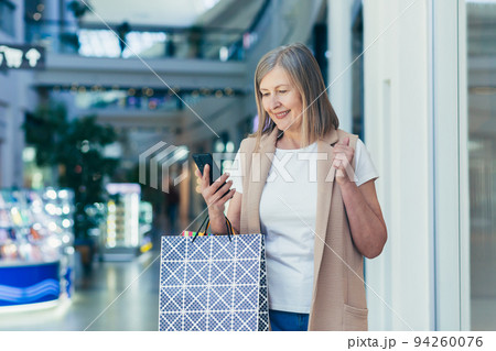 Portrait of a beautiful senior woman shopping in the mall. She uses the phone, makes purchases online, won a gift, rejoices. Holds paper shopping bags in her hands. She smiles, happy with her shopping 94260076