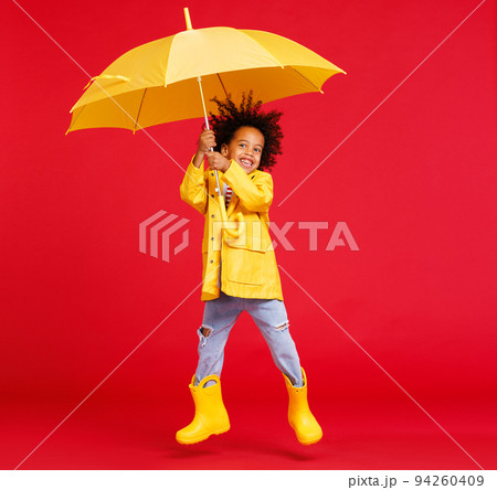 Cheerful ethnic boy jumping with umbrella against red backdrop Cheerful ethnic boy jumping with umbrella against red backdrop 94260409
