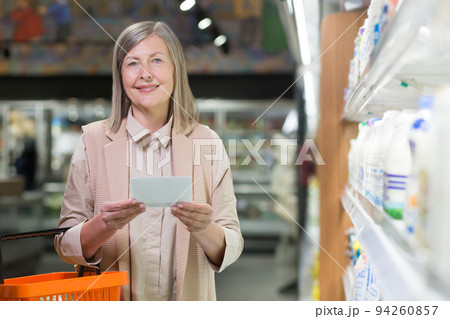 Portrait. Senior beautiful woman shopping in the store. Supermarket. Stands, holds a list of products written on paper in his hands, holds a plastic basket for products, chooses. He looks at the Portrait. Senior beautiful woman shopping in the store. Supermarket. Stands, holds a list of products written on paper in his hands, holds a plastic basket for products, chooses. He looks at the 94260857