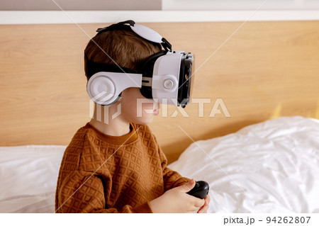 Little adorable boy sitting on bed at home with VR headset and playing interactive video game, exploring virtual reality. Cute child wearing VR glasses. Future, gadgets, technology, education online. 94262807