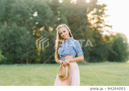 A beautiful young girl in light clothes holds a hat in her hands while walking in a clearing in the forest 94264001
