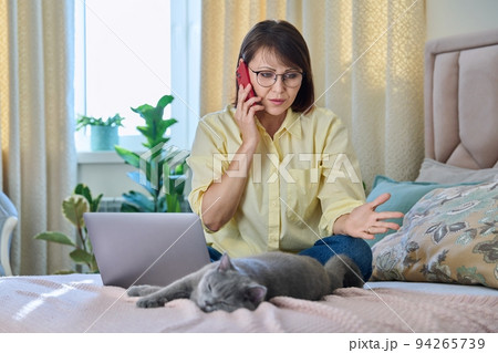 Middle-aged woman sitting on bed with laptop, cat talking on phone Middle-aged woman sitting on bed with laptop, cat talking on phone 94265739