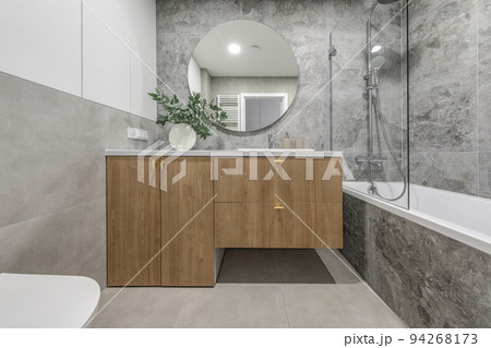 Modern minimalist bathroom interior design with wooden furniture, grey stone tiles, round mirror. Aesthetic simple interior design concept. Modern minimalist bathroom interior design with wooden furniture, grey stone tiles, round mirror. Aesthetic simple interior design concept. 94268173