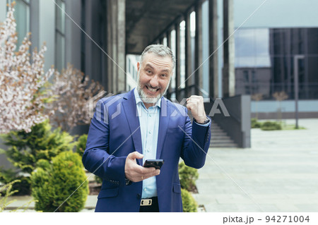 A senior gray-haired man in a business suit is standing near an office center with a phone in his hands. Rejoices, celebrates winning the lottery, plays on the phone. Shows with his hand yes. 94271004