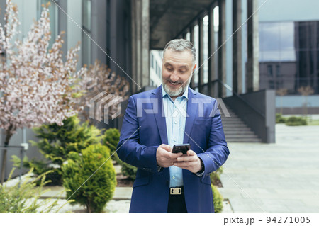 A senior gray-haired man in a business suit is standing near an office center with a phone in his hands. Rejoices, celebrates winning the lottery, plays on the phone. 94271005
