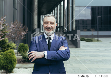 Portrait of a successful senior handsome man, businessman, judge, lawyer. Standing in a suit near the courthouse, office center. Crossed his arms in front, looks at the camera, smiles. 94271006