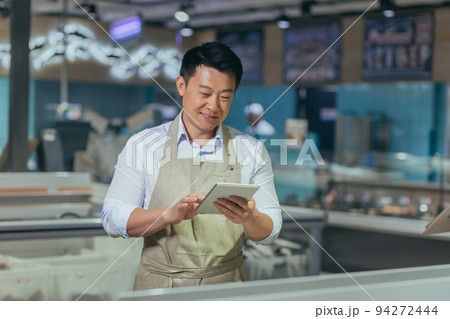 Asian grocery store manager salesman in apron using digital tablet counting goods in supermarket worker employee doing inspection of products regular inventory to control goods online order 94272444