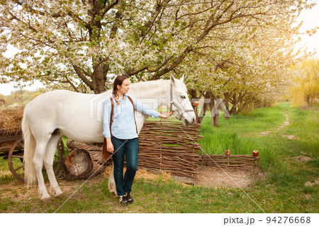 A beautiful young woman poses next to a white horse. Cowboy, ranch, village. Love friendship concern. Pet. Outside the door, green grass woods. sunlight. Blurred background, screensaver magic A beautiful young woman poses next to a white horse. Cowboy, ranch, village. Love friendship concern. Pet. Outside the door, green grass woods. sunlight. Blurred background, screensaver magic 94276668