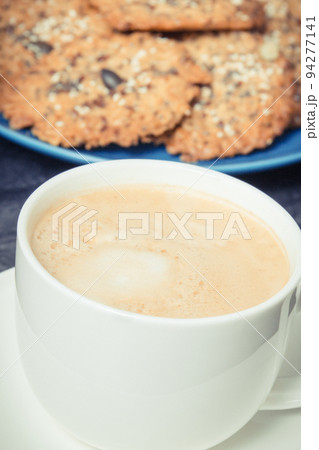 Cup of white coffee and fresh baked oatmeal cookies with honey and healthy seeds on blue plate Cup of white coffee and fresh baked oatmeal cookies with honey and healthy seeds on blue plate 94277141