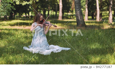 A young girl plays the violin on the sitting grass in the park. A young girl plays the violin on the sitting grass in the park. 94277187