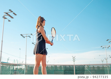 A beautiful young woman plays tennis on the court. Sporting a black suit dress. Blue sky, empty space for text. The girl is giving the ball. Exercise, active lifestyle, concept. portrait. back view 94277335